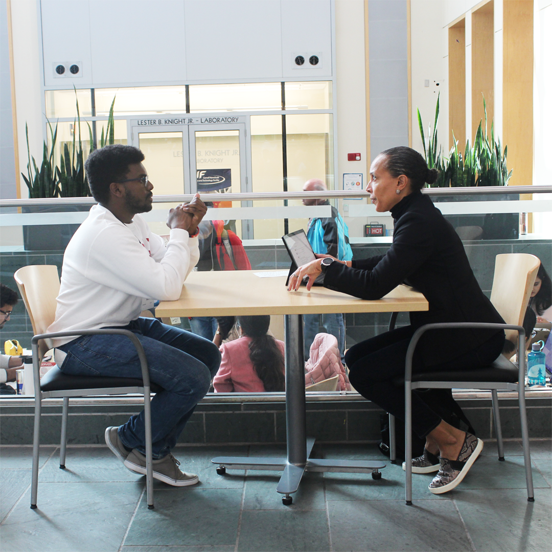 A color photo of two people sitting at a table, speaking with each other